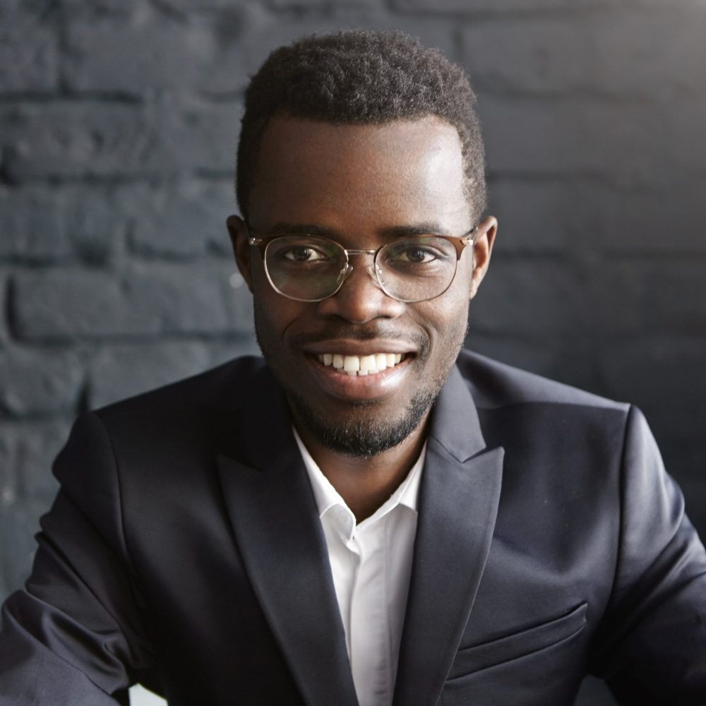 Portrait of confident and successful young African-American businessman wearing stylish glasses and elegant dark formal suit smiling happily, rejoicing at profitable deal he has just concluded